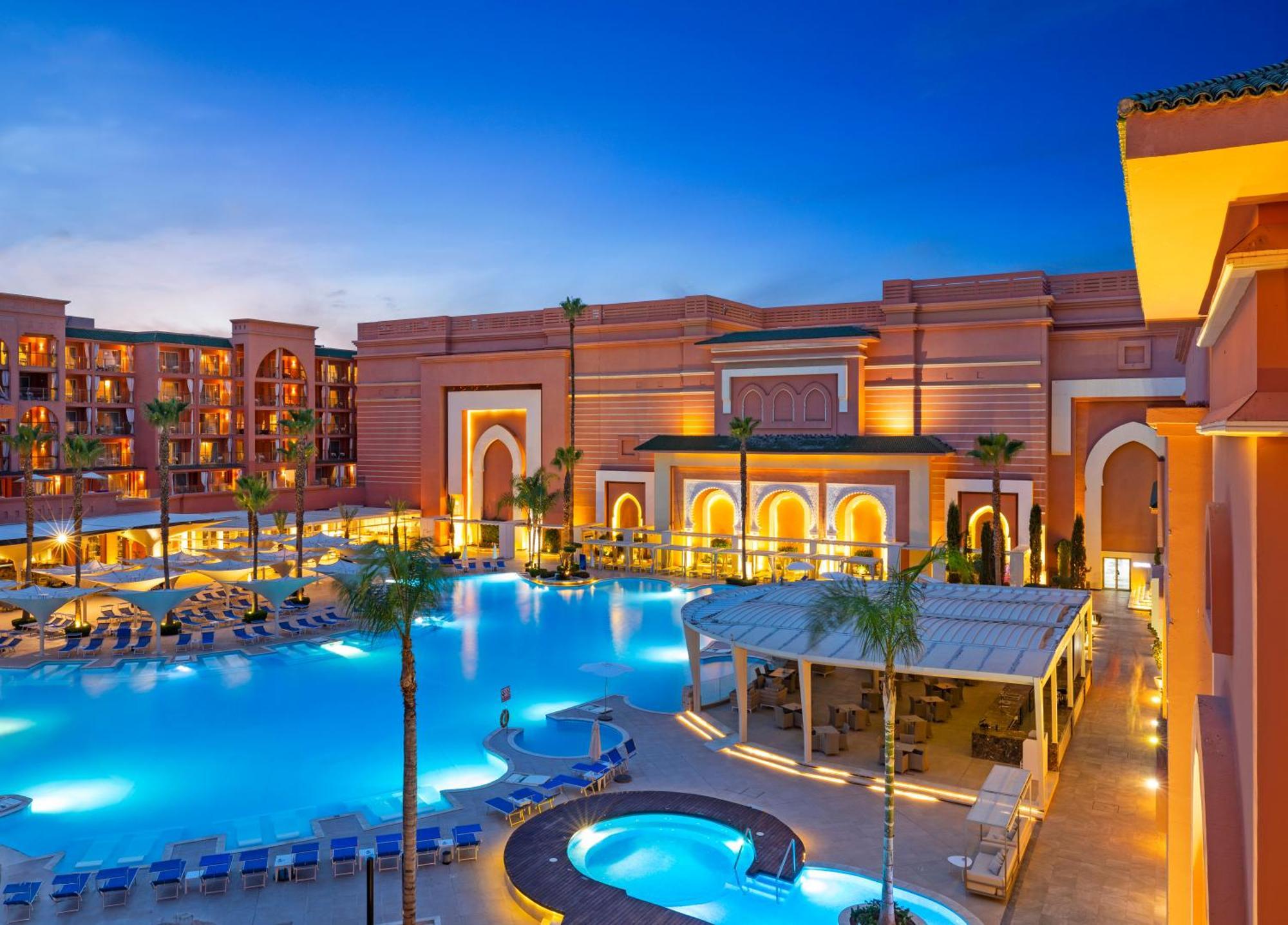 Savoy Le Grand Hotel Marrakech Marrakesh Exterior photo The photo depicts a luxurious hotel or resort setting, showcasing a large swimming pool that is illuminated in a vibrant blue color. Surrounding the pool are lounge chairs, palm trees, and a gazebo or bar area. The architecture features a rich and in