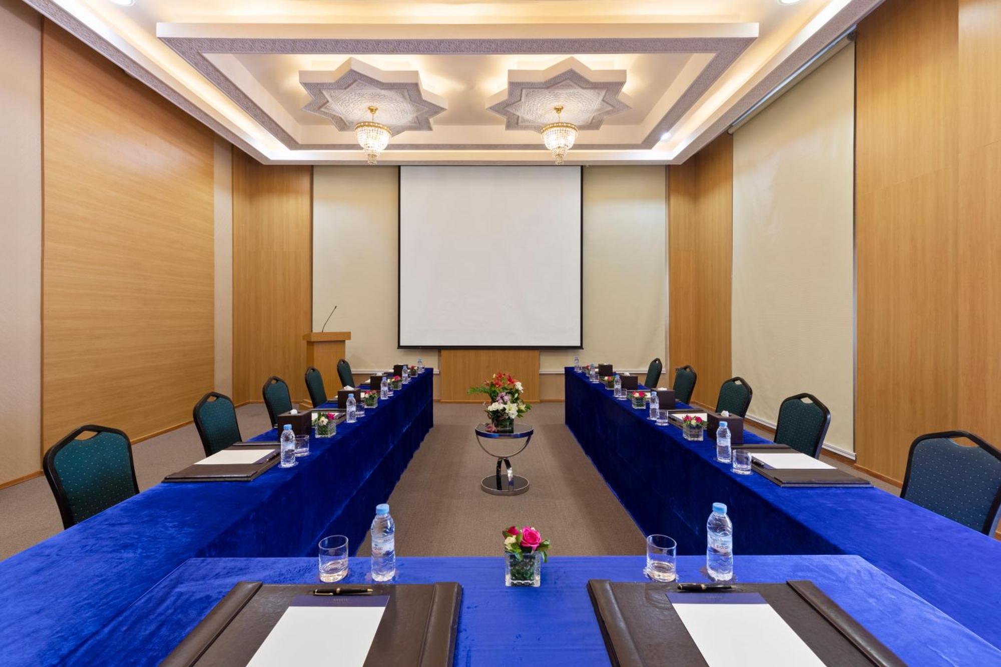 Savoy Le Grand Hotel Marrakech Marrakesh Exterior photo The image depicts a conference room set up for a meeting. The room features a long table covered with blue tablecloths, with black folders and drinking water bottles placed at each seat. There are green chairs arranged around the table. At the front,